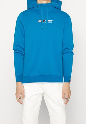 Niebieska bluza z kapturem Tommy Hilfiger z logo na piersi, noszona przez osobę w białych spodniach i srebrnych pierścieniach na palcach.
