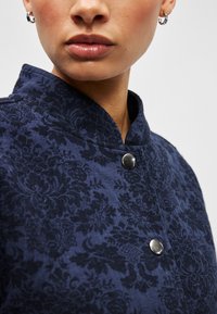 Μπλε ναυτικό υφασμάτινο σακάκι με λουλουδάτο μοτίβο, όρθιο γιακά και μεταλλικά κουμπιά στο μπροστινό μέρος.