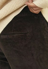 Pantalon en velours côtelé marron avec des côtes verticales, doté d'une poche latérale, porté sous un pull en tricot crème. Texture douce visible.