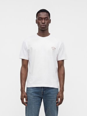 Homme portant un t-shirt blanc à manches courtes avec un petit texte rouge sur la poitrine et un jean bleu, debout devant un fond uni clair.