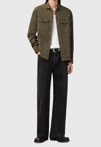 Olivgrüne Hemdjacke mit zwei Brusttaschen, kombiniert mit schwarzen, weit geschnittenen Jeans und schwarzen Stiefeletten. Weißes Unterhemd sichtbar.