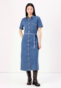 Modra denim oblečna srajca s kratkimi rokavi, z zapiranjem na gumbe, sprednjim žepom in tkanim pasom, skombinirana z rjavimi škornji do kolen.