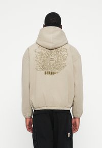Beige hættebluse med elastiske манchetter og kant. Har et grafisk design på ryggen, tekst "HVOR EN KAOS!" over den mørkegrå "HUF" logo.