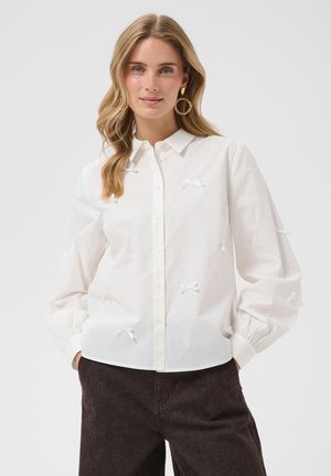 Femme aux cheveux blonds lâchés portant une chemise blanche boutonnée avec de petits nœuds, un pantalon sombre et de grandes boucles d’oreilles, debout devant un fond uni.