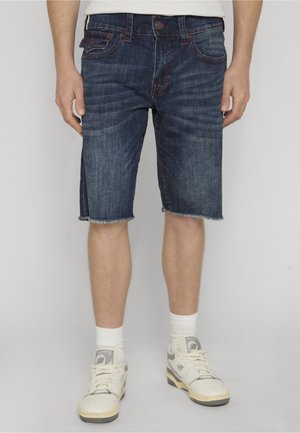 Osoba nosząca ciemnoniebieskie dżinsowe szorty z postrzępionymi brzegami, białe skarpety i białe tenisówki, stojąca na jednolitym tle.