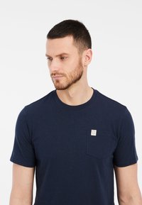 Námornícky modré tričko s krátkym rukávom vyrobené z mäkkej látky, s náprsné vreckom a štítkom, okrúhlym výstrihom a voľným strihom.