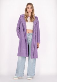 Cappotto viola soffice con cappuccio e grandi bottoni, abbinato a jeans a gamba larga blu chiaro e sneakers. Texture morbida e vestibilità rilassata.
