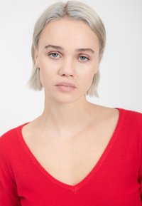Roter, taillierter Pullover mit V-Ausschnitt, gefertigt aus einem weichen, strukturierten Strick. Das Model hat kurzes, hellgraues Haar und einen neutralen Hintergrund.
