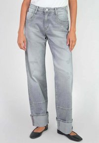 Jeans en denim gris clair avec une coupe ample, présentant des coutures visibles, des détails usés et des ourlets retroussés. Portés avec des chaussures noires plates.