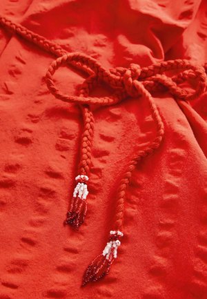 Roter Stoff mit einer strukturierten Oberfläche. Verfügt über ein geflochtenes Band mit beadedenden in Rot und Weiß. Der Stoff ist glatt und flexibel.