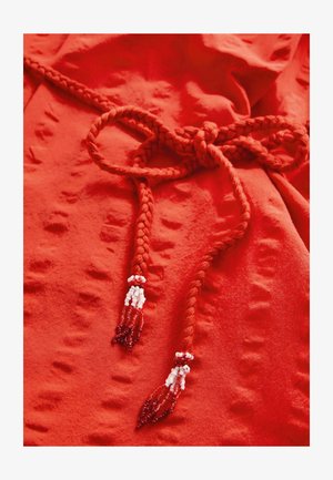 Roter Stoff mit einer strukturierten Oberfläche. Verfügt über ein geflochtenes Band mit beadedenden in Rot und Weiß. Der Stoff ist glatt und flexibel.