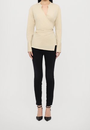 Kobieta ubrana w beżową, długą bluzkę kopertową, czarne dopasowane spodnie oraz czarne szpilki z paskiem wokół kostki na jednolitym tle.