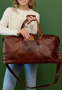 Ruskea nahkainen duffel-laukku kiiltävällä pinnalla, kaksinkertaiset kahvat, säädettävä olkahihna ja vetoketjutasku. Esillä myös osittain näkyvä lompakko.