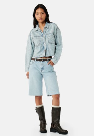 Kvinde iført en lyseblå cropped denimjakke med små prikker, knælange denimshorts, mørkebrune støvler og et brunt bælte.