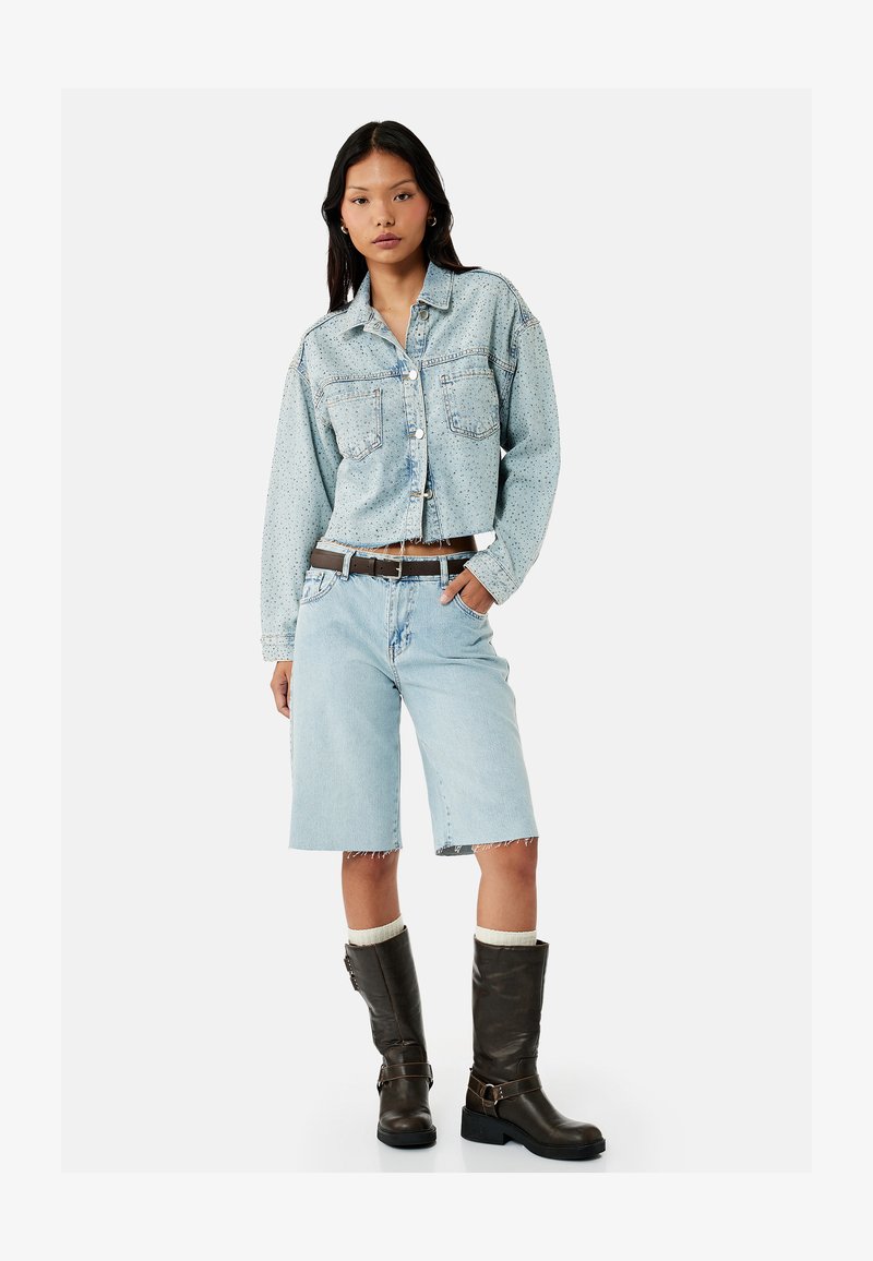 Kvinde iført en lyseblå cropped denimjakke med små prikker, knælange denimshorts, mørkebrune støvler og et brunt bælte.