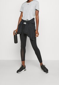 Camiseta deportiva gris con un pequeño logotipo negro, leggings negros con paneles de malla y zapatillas negras, sosteniendo una botella de agua negra.