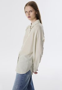 Crème-kleurig, loose-fit shirt met een knoopsluiting aan de voorkant, lange mouwen en een subtiele textuurpatroon, gedragen met lichtblauwe, versleten jeans.