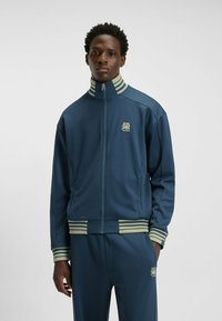 Marineblå zip-up træningsjakke med grønne striber på kraven og manchetterne, med et logo på venstre bryst og et glat, atletisk materiale.