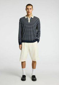 Polo a righe blu navy e bianche con maniche lunghe e colletto beige, abbinato a pantaloni corti color crema, calzini bianchi e scarpe nere.