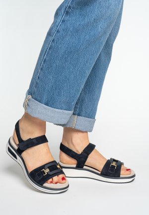 Feet wearing black platform sandals with gold buckles and blue rolled-up jeans against a white background.