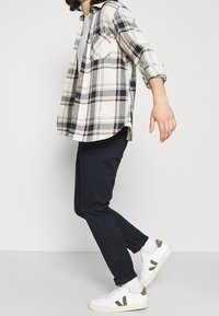 Camicia di flanella a quadri in crema, blu navy e pesco, abbinata a pantaloni scuri e sneakers bianche. La camicia presenta maniche arrotolate e un dettaglio sulla tasca.