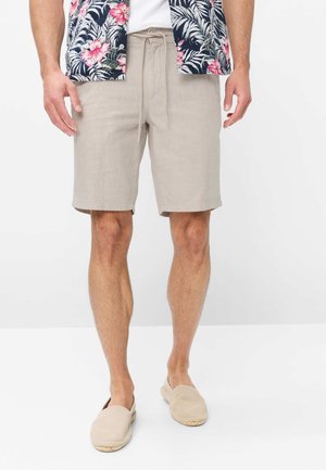 Mann trägt beige Shorts mit Kordelzug, beige Slip-On-Schuhe und ein kurzärmliges, geblümtes Hemd mit rosa Blumen und grünen Blättern.
