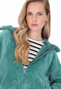 Türkisfarbene Fleecejacke mit Reißverschluss und zwei seitlichen Taschen sowie strukturierter Oberfläche, getragen über einem schwarz-weiß gestreiften Hemd.