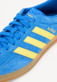 Blå ruskinds-sneakers med tre gule striber, teksturerede snørebånd og en gummirigtig sål. Features "GA ZELLE" branding på siden.