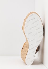Semelle d'une sandale compensée beige avec côtés tissés, semelle blanche embossée d'un texte répété "TOMMY JEANS", sur un fond blanc en coin.
