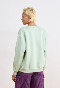 Sudadera oversized verde menta con cuello redondo y puños acanalados, combinada con pantalones cargo morados. Textura suave, corte relajado.