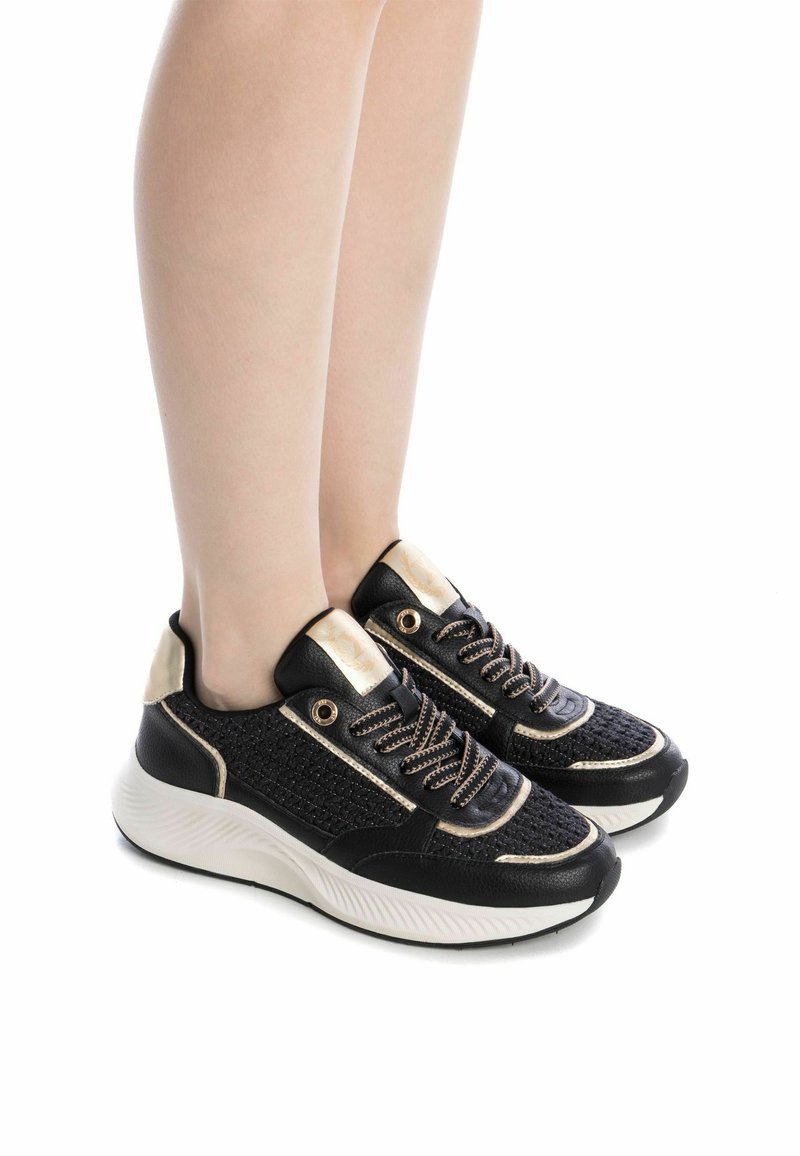 Zapatillas negras y doradas con parte superior tejida, entresuela blanca redondeada y cordones con acentos metálicos. Materiales texturizados y diseño estiloso.