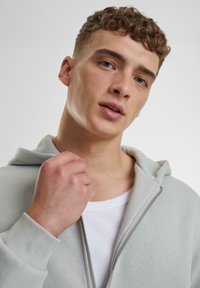 Hellgrauer Reißverschluss-Hoodie mit weicher Textur und geräumiger Kapuze, getragen über einem weißen gerippten Tanktop. Mit sichtbarem Reißverschluss und Nähdetails.