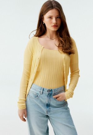 Geel geribbelde cardigan met knopen, gecombineerd met een bijpassend geribbeld tanktop en lichtblauwe wijde jeans, met een relaxte pasvorm.