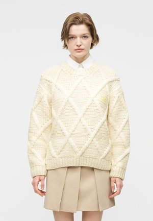 Νεαρό άτομο με κοντά μαλλιά που φοράει κρεμ χοντρό πλεκτό πουλόβερ πάνω από λευκό πουκάμισο με γιακά και μπεζ πλισέ φούστα.