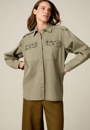 Vrouw draagt losse olijfgroene blouse met gestudeerde borstzakken en bruine wijde broek, poseert met handen in haar zij.