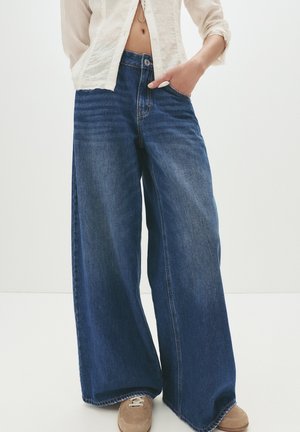 Wijduitlopende denim jeans in donkerblauw met een raw hem, voorzien van voorzakken en subtiele vervaging. Gecombineerd met beige sneakers.