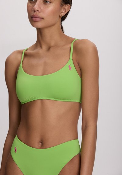 Φωτεινή πράσινη μπικίνι κορυφή με λεπτές τιράντες, διακοσμημένη με ροζ λογότυπο. Το υλικό είναι λείο και ελαστικό, σχεδιασμένο για άνεση και εφαρμογή.