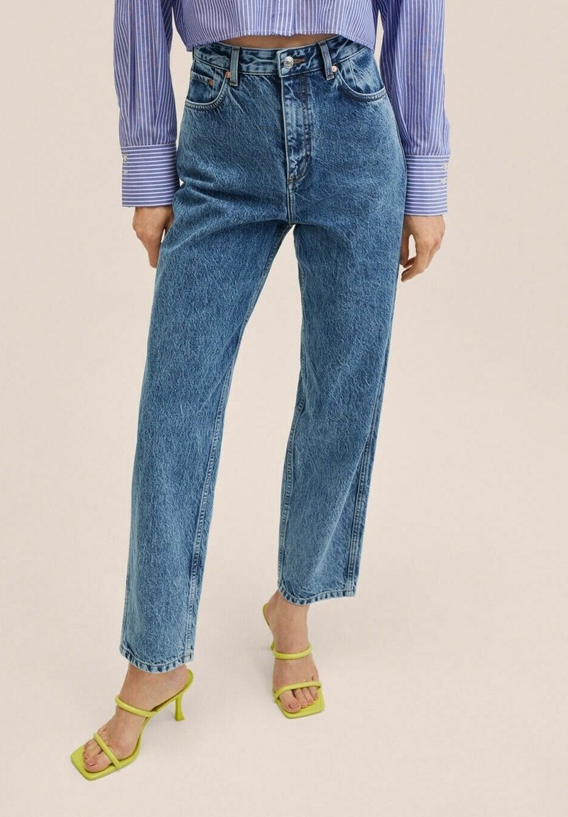 Jeans de mezclilla azul de cintura alta con un diseño de pierna recta, con un lavado descolorido y acentuados por sandalias de tacón alto verdes con tiras.