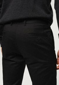 Schwarze Hosen mit tailliertem Schnitt, glatter Textur und einer Gesäßtasche mit Knopfdetail. Kombiniert mit einem dunklen Pullover.