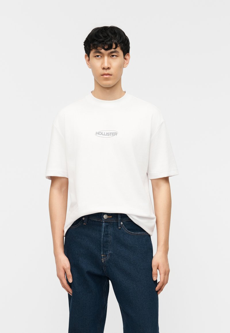 Νεαρός άνδρας με κοντά μαύρα μαλλιά, φορούσε λευκό T-shirt Hollister και μπλε τζιν, στέκεται μπροστά σε απλό ανοιχτόχρωμο φόντο.