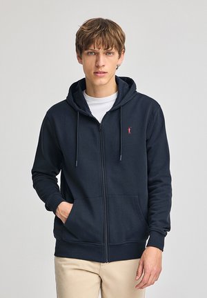 Hoodie zippé bleu marine en tissu doux, doté d'une poche avant, d'une capuche avec cordon de serrage et d'un petit logo rouge sur la poitrine à gauche.