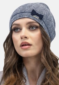 Gorra de punto en patrón de damero en azul marino y gris, con un acento de lazo de fieltro y un pequeño detalle de logotipo metálico en el lado.