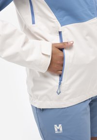 Leichte, zweifarbige Jacke mit einem blauen oberen Teil und einem weißen unteren Teil, ausgestattet mit einer Reißverschlusstasche und verstellbaren Bündchen, hergestellt aus einem strukturierten Material.