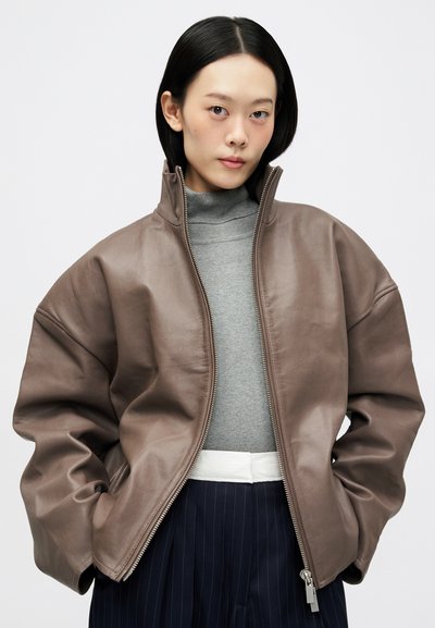 Νεαρός με ίσια μαύρα μαλλιά φοράει oversized καφέ φερμουάρ, γκρι τρύπα λαιμού και σκούρες ριγέ παντελόνες σε έναν απλό φόντο.