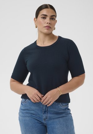 Femme aux cheveux foncés en chignon bas, portant des boucles d'oreilles créoles en or, un haut bleu marine à manches courtes, un jean bleu et un bracelet en or, debout les mains jointes.