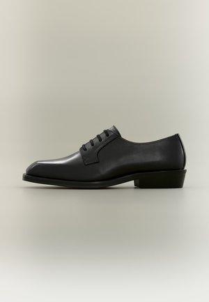 Chaussure habillée pour homme en cuir noir avec laçage fermé, talon bas et bout arrondi sur un fond neutre.