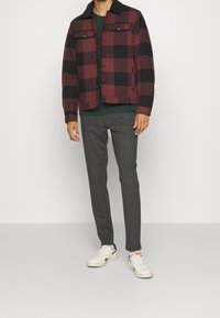 Czerwona i czarna flanelowa kurtka w kratę z ciemnym kołnierzem, zielona koszula, szare spodnie i białe trampki z bordowymi akcentami.