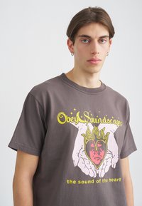 Camiseta de algodón gris oscuro con un gráfico colorido que muestra manos sosteniendo un corazón, llamas y el texto: "Obedece Paisajes Sonoros" y "el sonido del corazón."