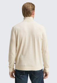 Beige gebreide pullover trui met een hoge kraag, lange mouwen en geribbelde manchetten. Zachte textuur, gecombineerd met donkerblauwe jeans.