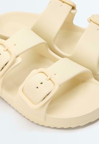 Beige Sandalen mit offenem Zehenbereich, zwei verstellbaren Schnallenriemen und einer strukturierten Sohle vor weißem Hintergrund.